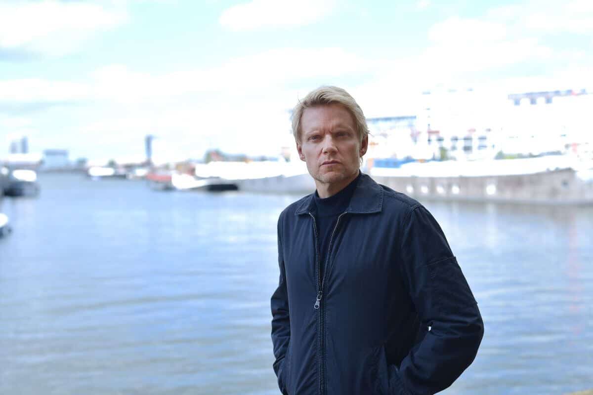 Marc Warren as Commissaris Piet Van der Valk with water and a boat behind him.